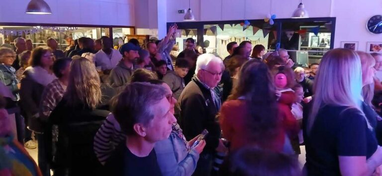 Foule de personnes réunies lors d'un événement festif en intérieur, avec guirlandes de fanions colorés et ambiance chaleureuse